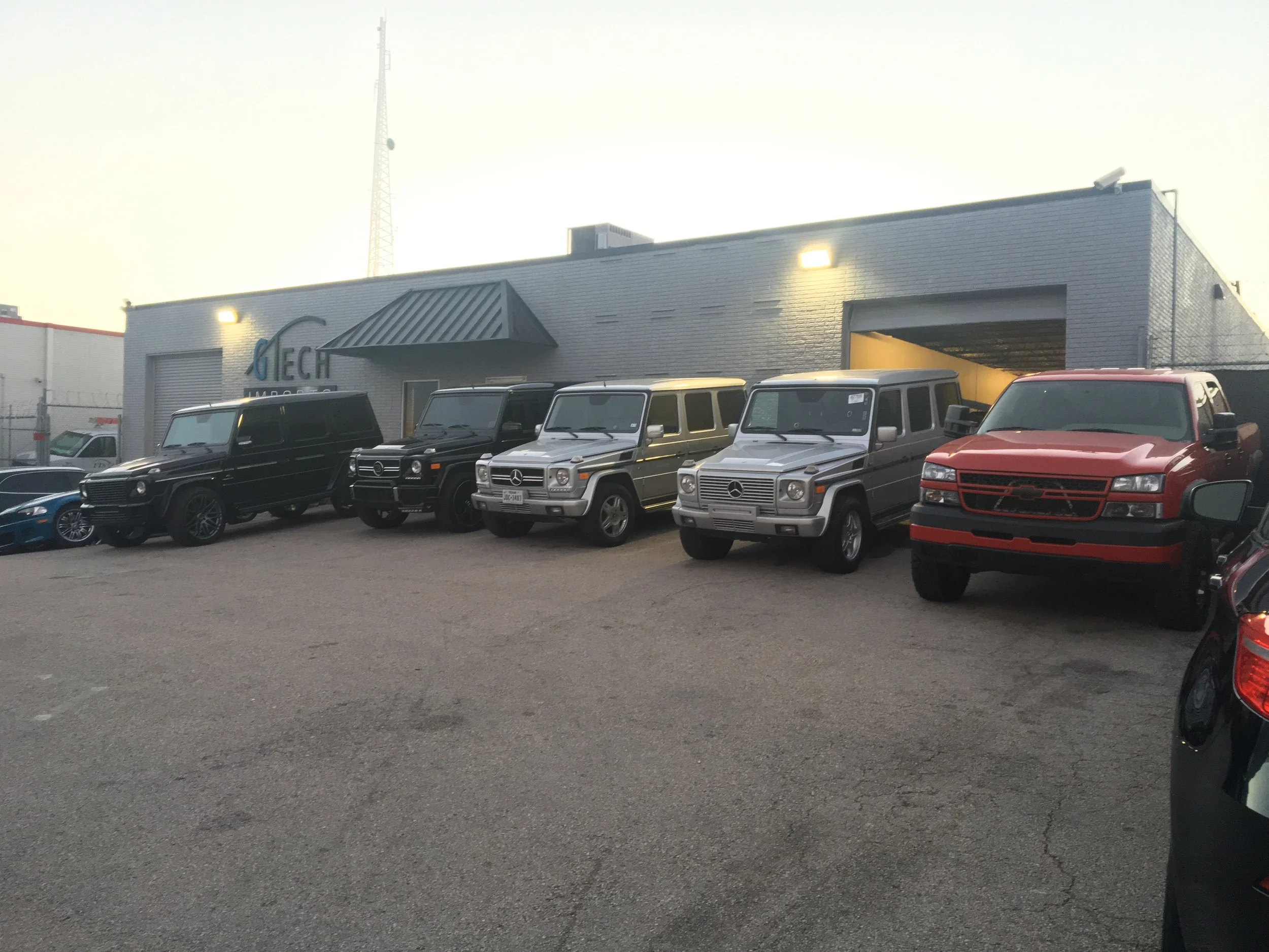 Line of luxury and classic SUVs, including Mercedes-Benz G-Class models and a Chevrolet Silverado truck, parked outside a grey industrial-style building with the partially visible sign reading 'TECH'.