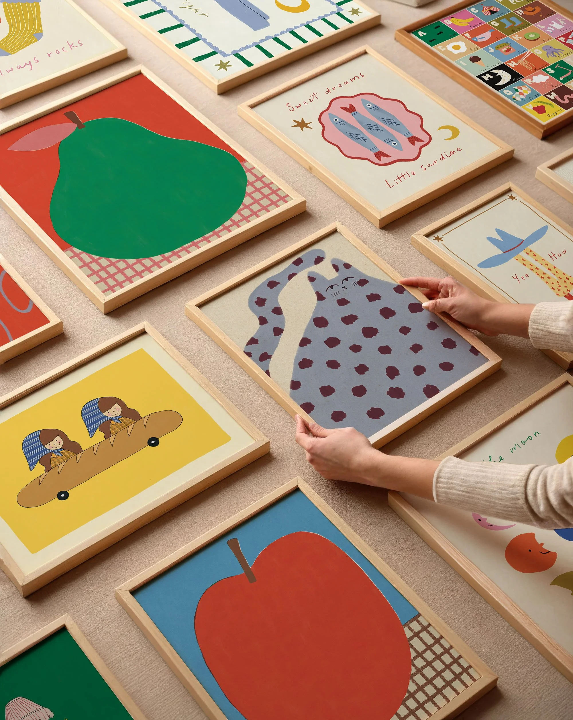 Collection of colorful framed illustrations for children laid out on a table, including a pear, a leopard, a red apple, a woman on a skateboard, and other playful images with text.