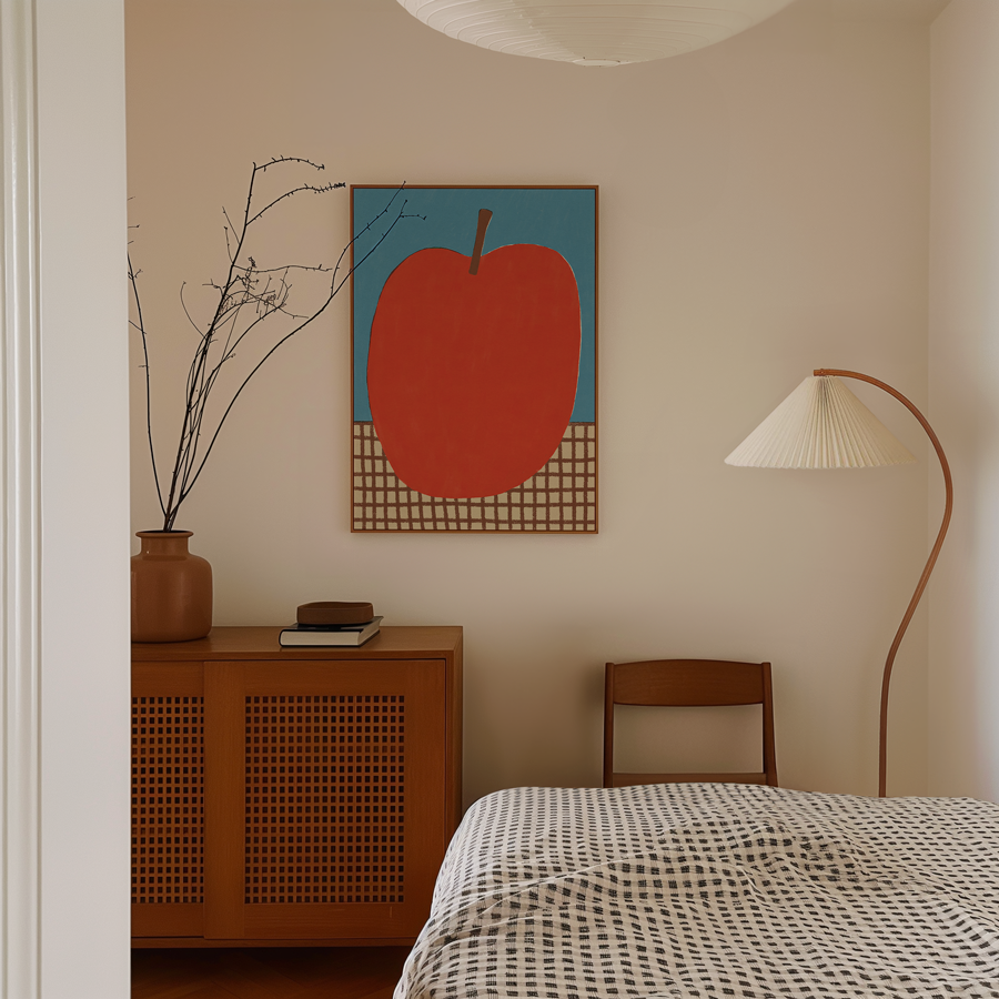 A minimalist bedroom with a framed pop art painting of a red apple on a blue background on the wall. There is a wooden sideboard with a brown vase holding dried branches, some books, and a small object. A wooden chair is next to a tall, curved floor lamp with a pleated shade. Part of a bed with a black and white patterned blanket is visible in the foreground.