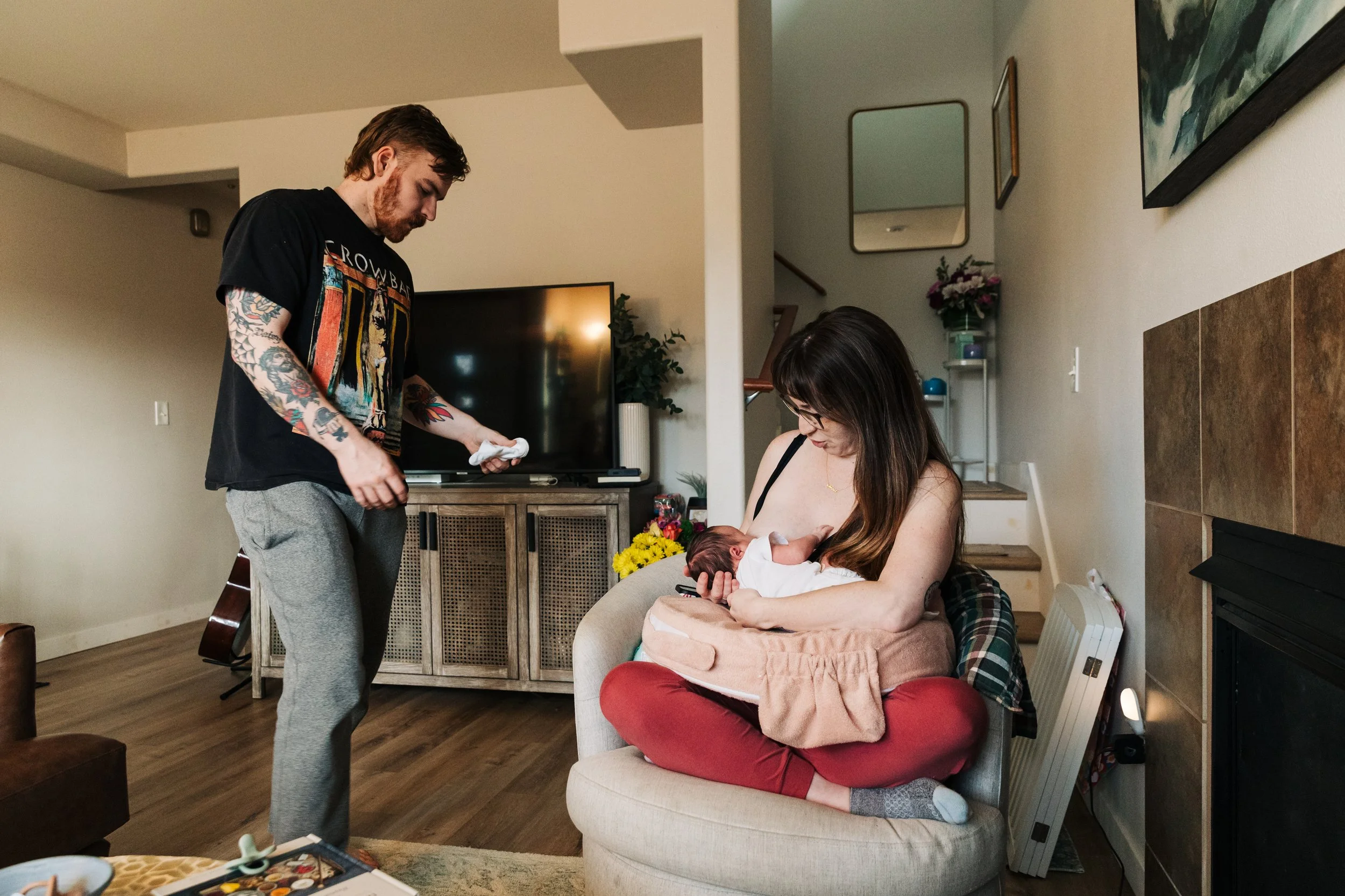 A mother nursing her newborn while a father hands them a burp cloth.