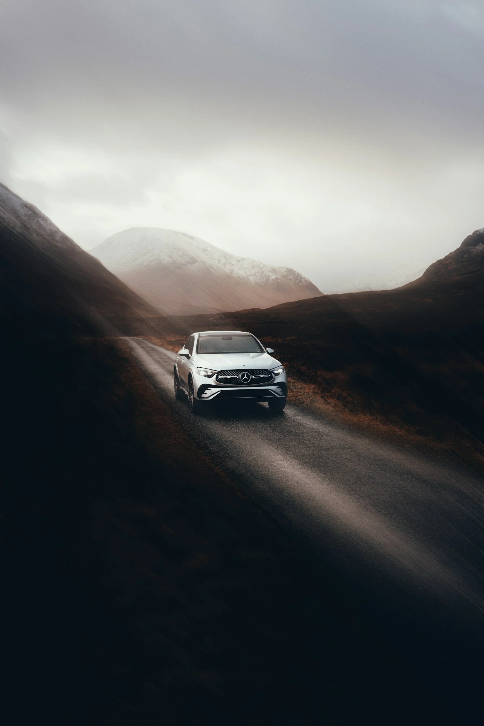 Silver Mercedes-Benz SUV driving on a winding mountain road under cloudy sky with snow-capped mountains in the background.