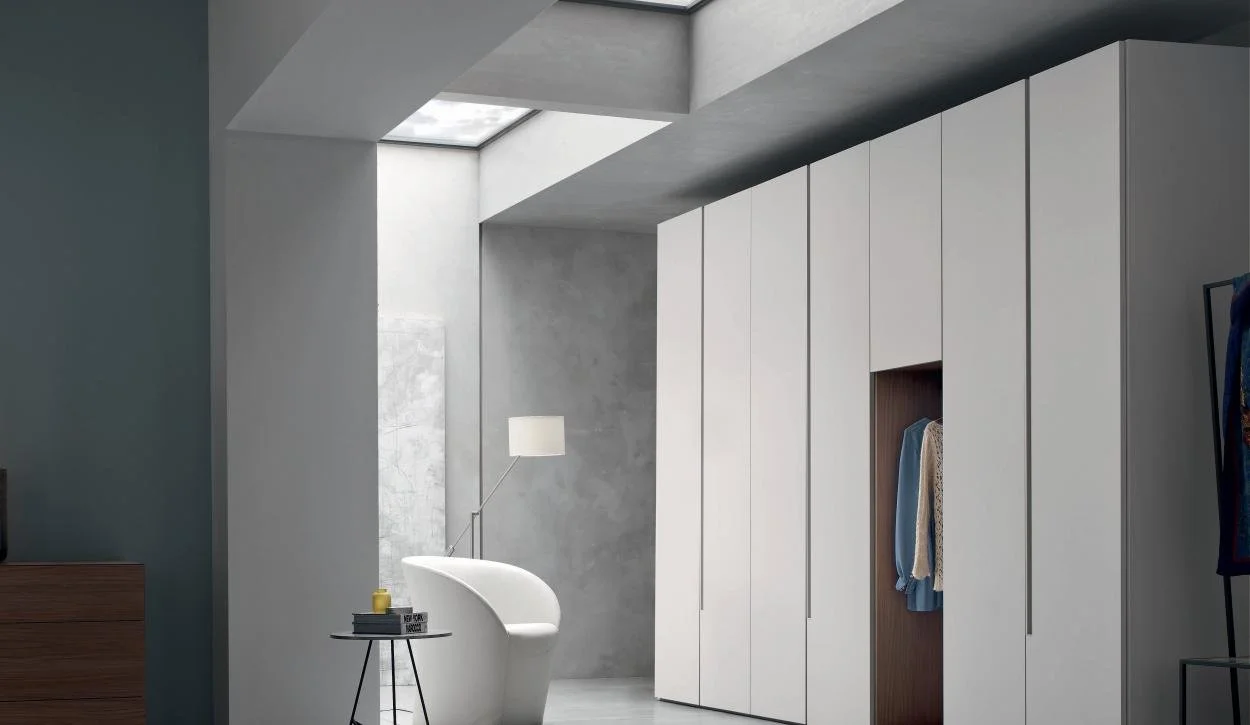 Modern minimalist bedroom with white built-in wardrobe, gray walls, a white lounge chair, and a small side table with a yellow candle and books, near a window with natural light.