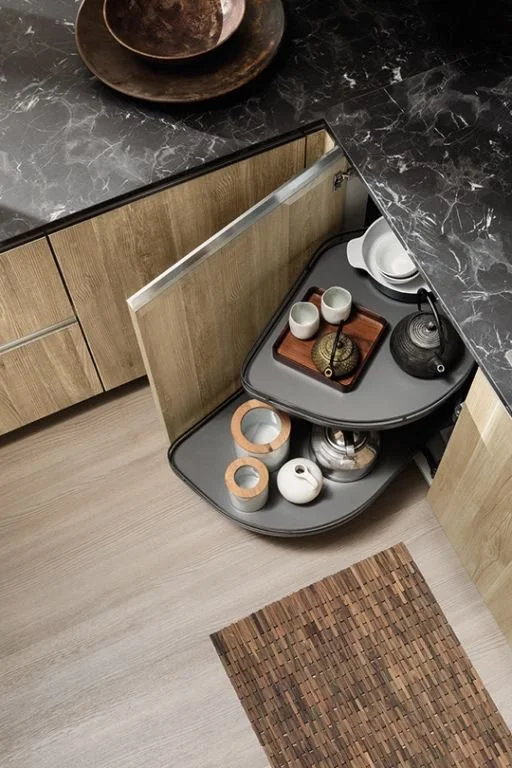 A corner of a kitchen with a wooden cabinet and black marble countertop, showing an open lazy Susan with various ceramic and decorative items, and a woven brown rug on light hardwood flooring.