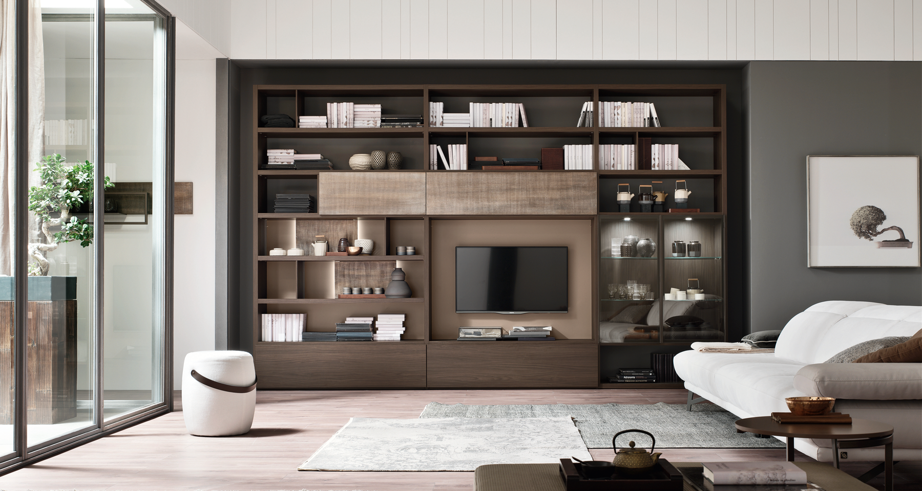 Modern living room with a large wooden bookshelf, flat-screen TV, white sofa, side tables, and a sliding glass door leading to a balcony with potted plants.