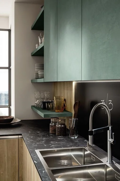 Close-up of a modern kitchen countertop with a black marble surface, a stainless steel sink, a curved faucet, and green cabinetry with open shelves holding dishes and glassware.