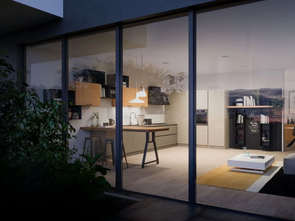 Photo of a modern kitchen and living room seen through a large glass window at dusk. The kitchen has beige cabinets, a wooden countertop, and bar stools, with pendant lighting above. The living room has a white coffee table, a yellow rug, and a black