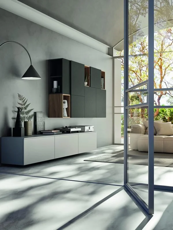 Modern living room with a white sofa, large glass windows showing trees outside, and minimalist decor including a black and wood wall-mounted shelf, a white cabinet, and a black hanging lamp, with sunlight casting shadows on the floor.