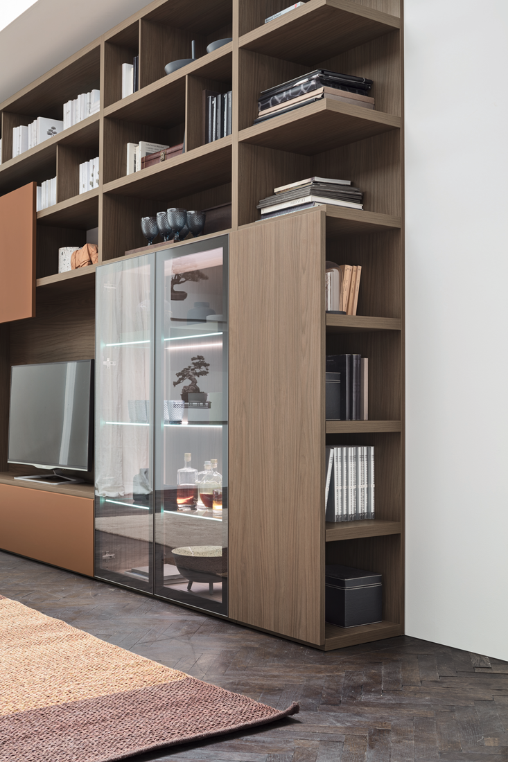 A modern wooden bookshelf with open shelves and a glass-front display cabinet, holding decorative items, books, and bottles, located in a living room with a flat-screen TV and a beige and brown rug.