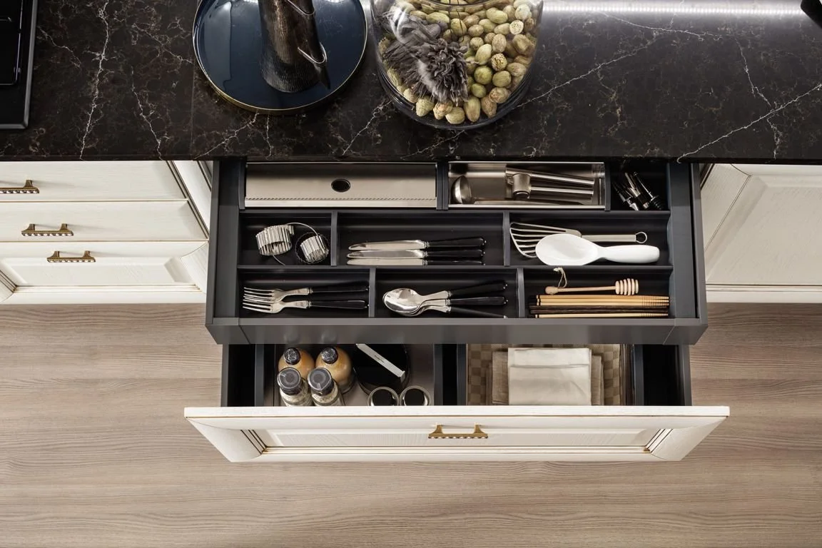Open kitchen drawer with utensils, including forks, knives, spoons, and a spatula, with a marble countertop above and white cabinetry.