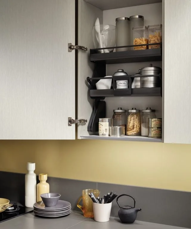 Kitchen cabinet opened to reveal shelves with jars of pasta, sugar, and other ingredients. Counter with bowls, utensils, bottles, and a teapot.