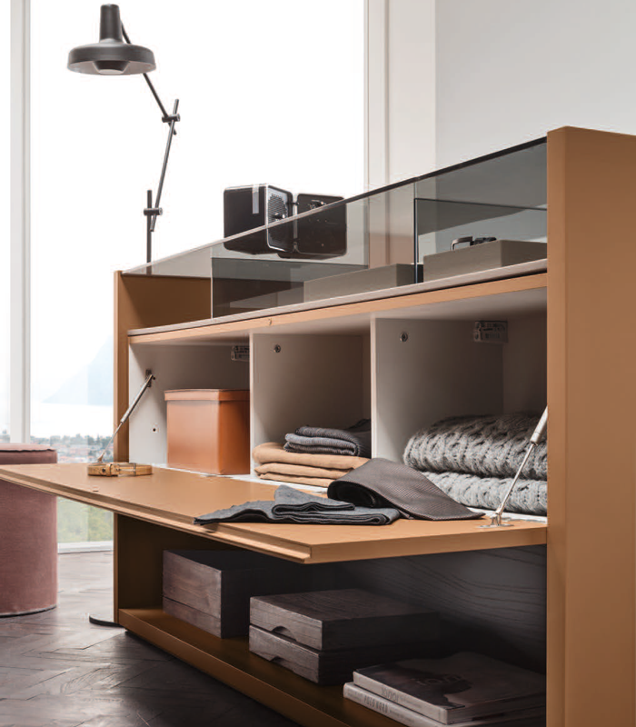 Modern desk with stacks of folded clothing, boxes, and electronic devices, next to a large window with a view of nature.