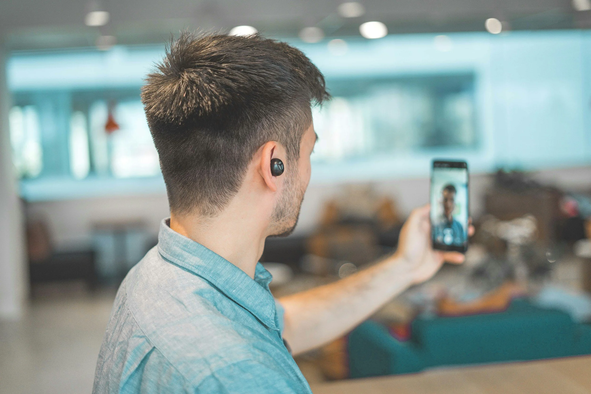 Man taking a selfie with smartphone in a modern indoor space, wearing wireless earbuds.