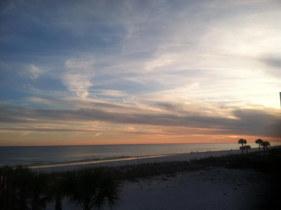 Sunset over the ocean with a sandy beach, palm trees, and a few people in the distance.