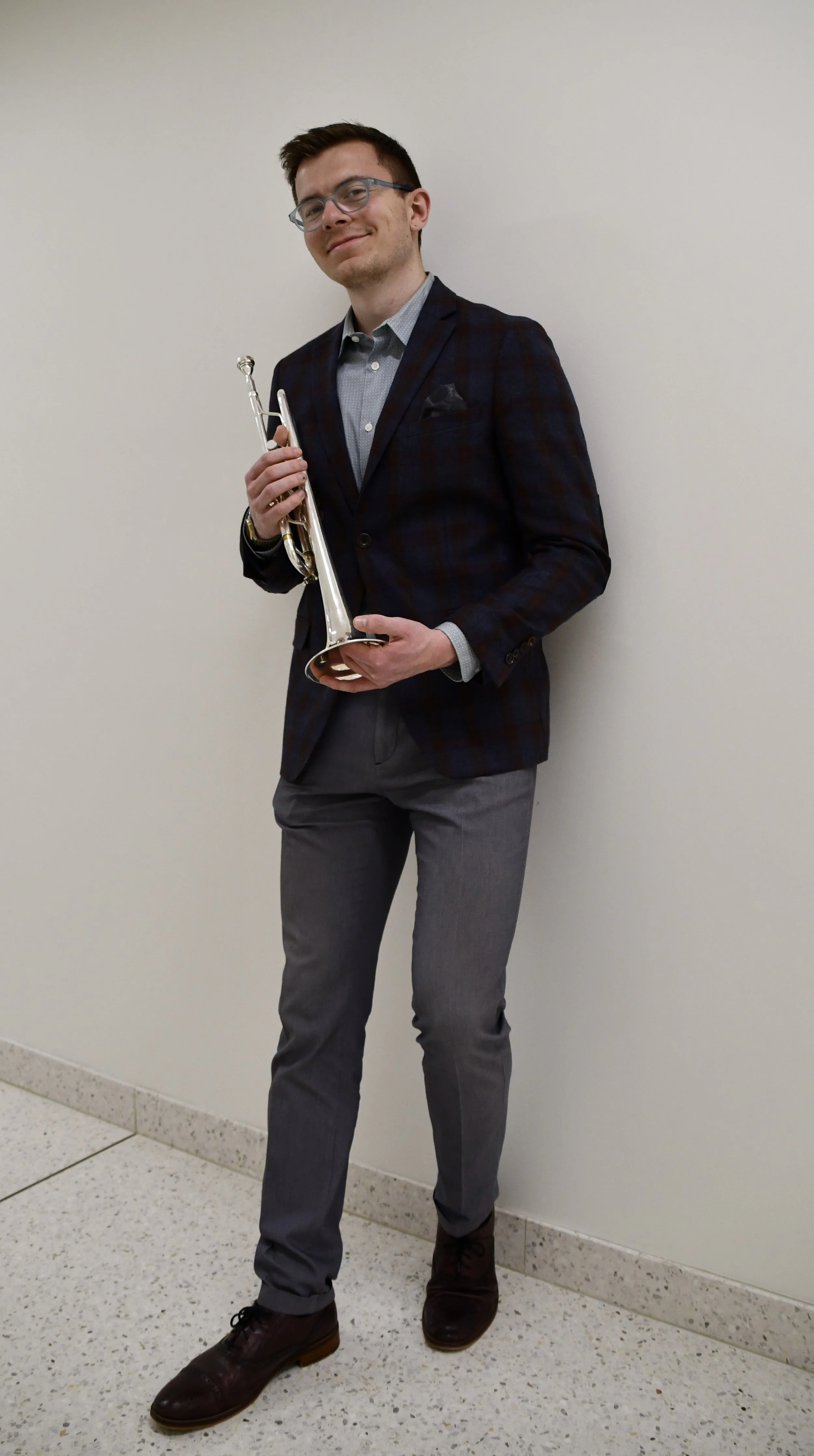 A young man in a dark plaid blazer, light shirt, and gray pants holding a silver trumpet, standing against a plain beige wall.
