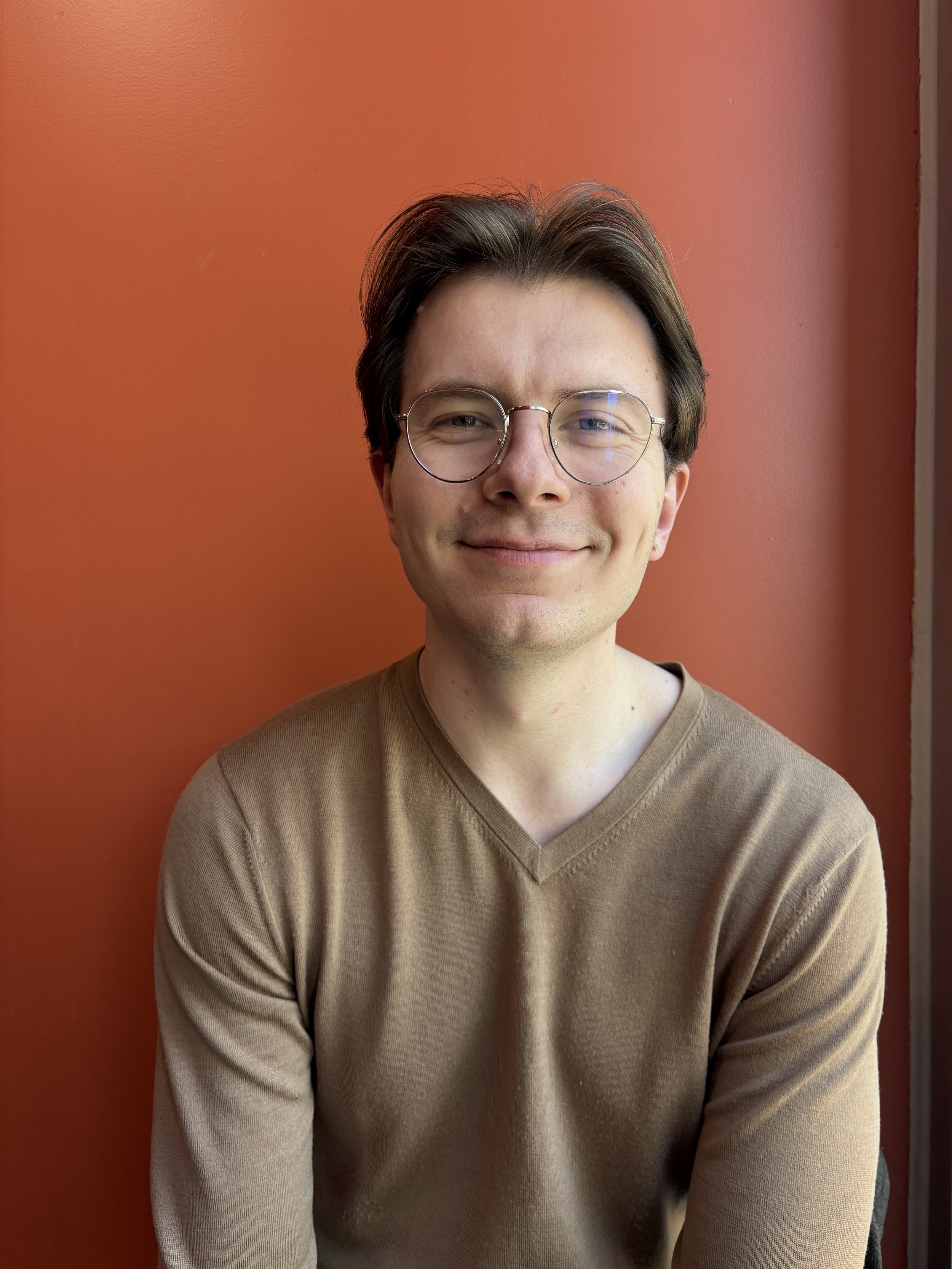A young man with dark hair, glasses, and light skin, smiling, standing in front of an orange wall, wearing a brown V-neck sweater.