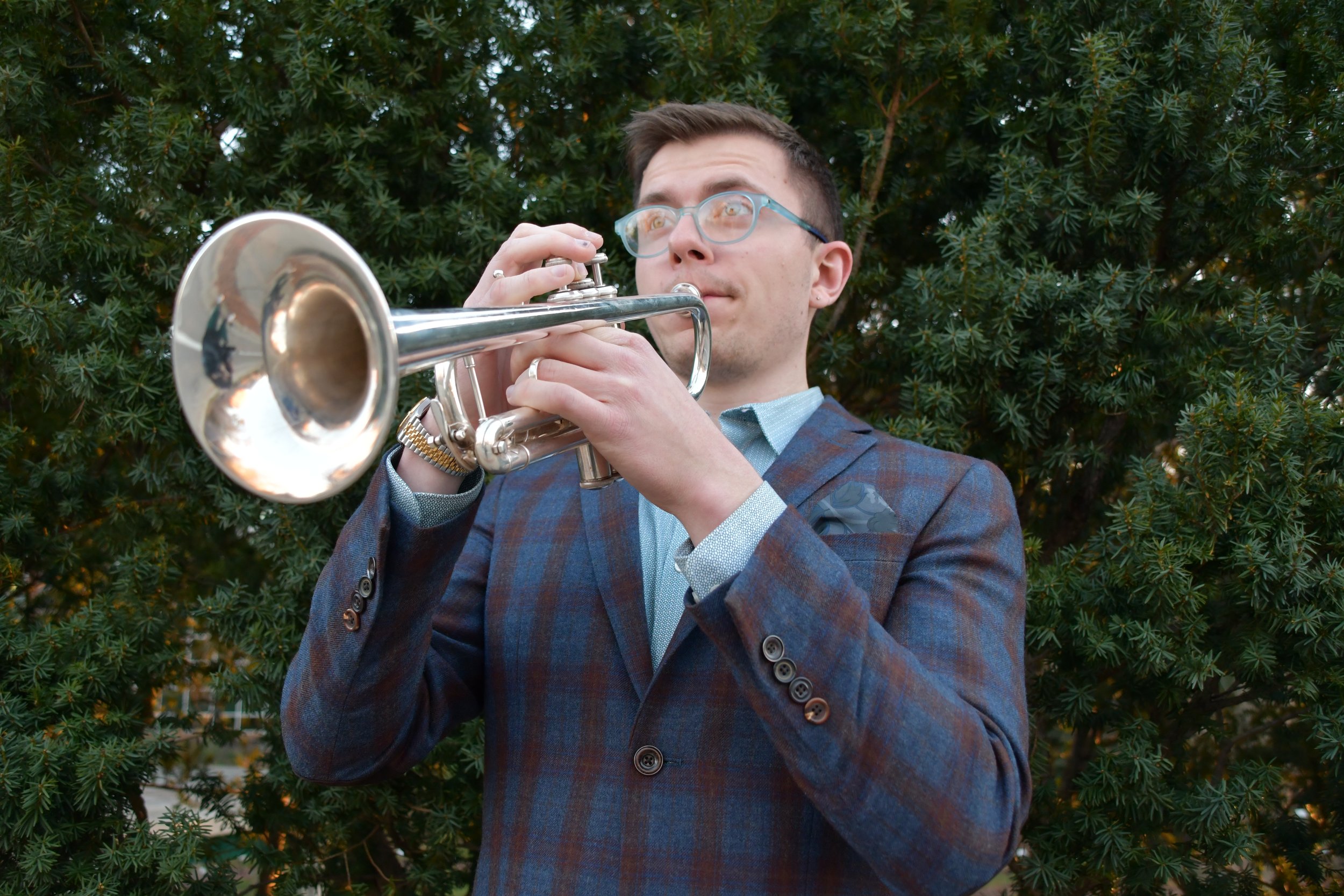 A man wearing a plaid blazer and glasses playing a trumpet outdoors with green bushes in the background.