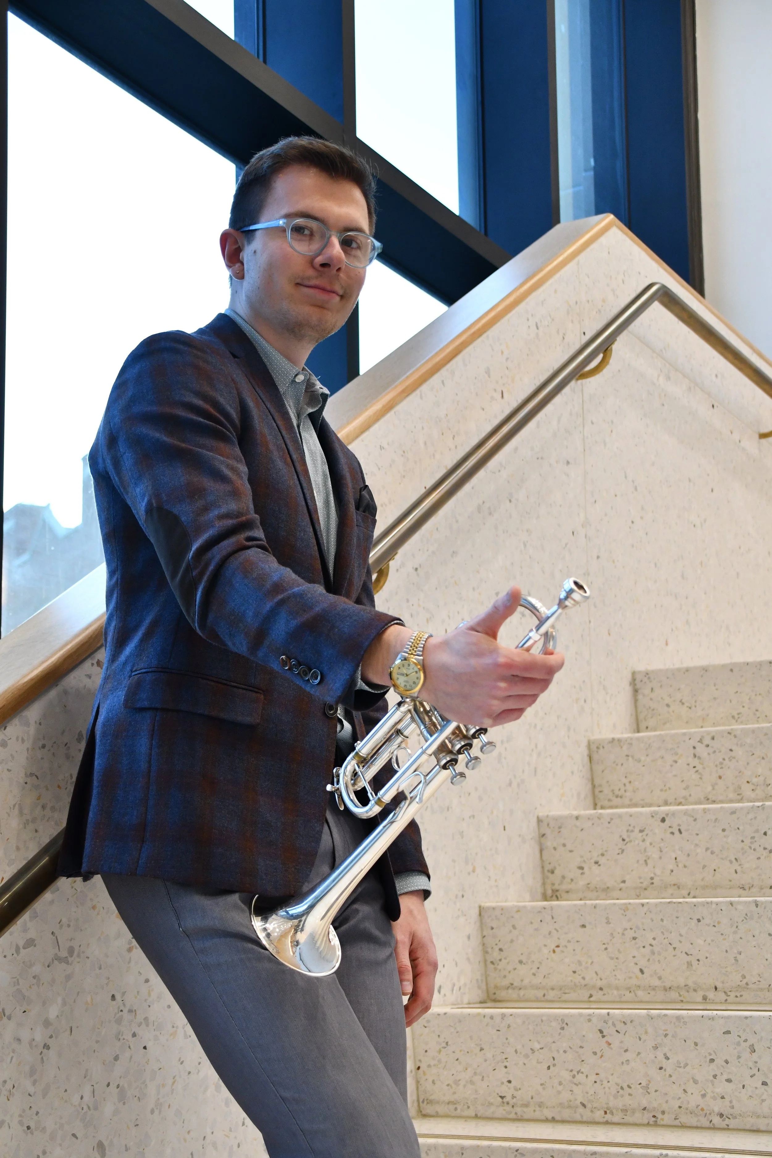 A man is standing on a staircase holding a silver trumpet and looking at the camera. He is wearing glasses, a wristwatch, a checkered blazer, a light-colored shirt, and gray pants. The staircase has a beige speckled surface with a metal handrail, and there are large windows in the background showing daylight.