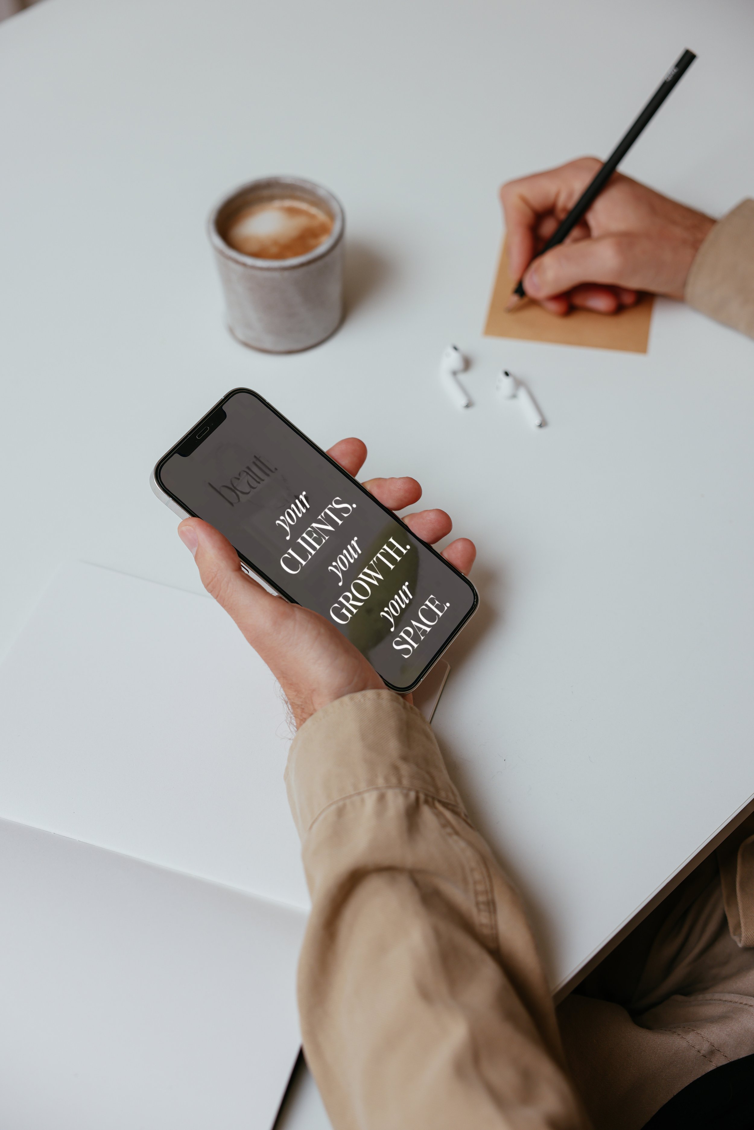 Person holding smartphone with the text 'your CLIENTS. your GROWTH. your SPACE.' on the screen, on a white desk with a coffee cup, a pair of wireless earbuds, and someone writing on a sticky note.