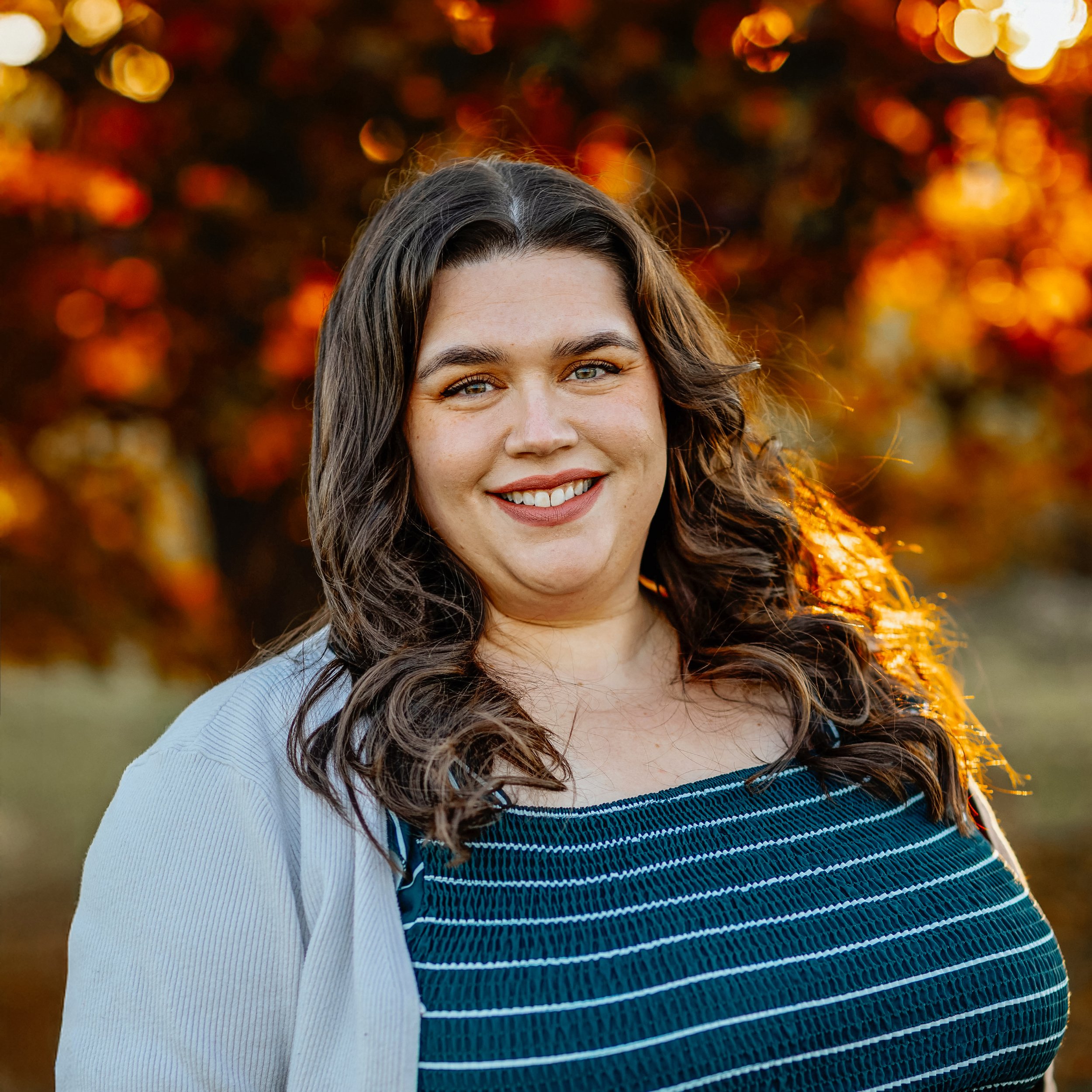 A woman with wavy brown hair and blue eyes smiling outdoors in front of a background of autumnal trees with orange and red leaves.