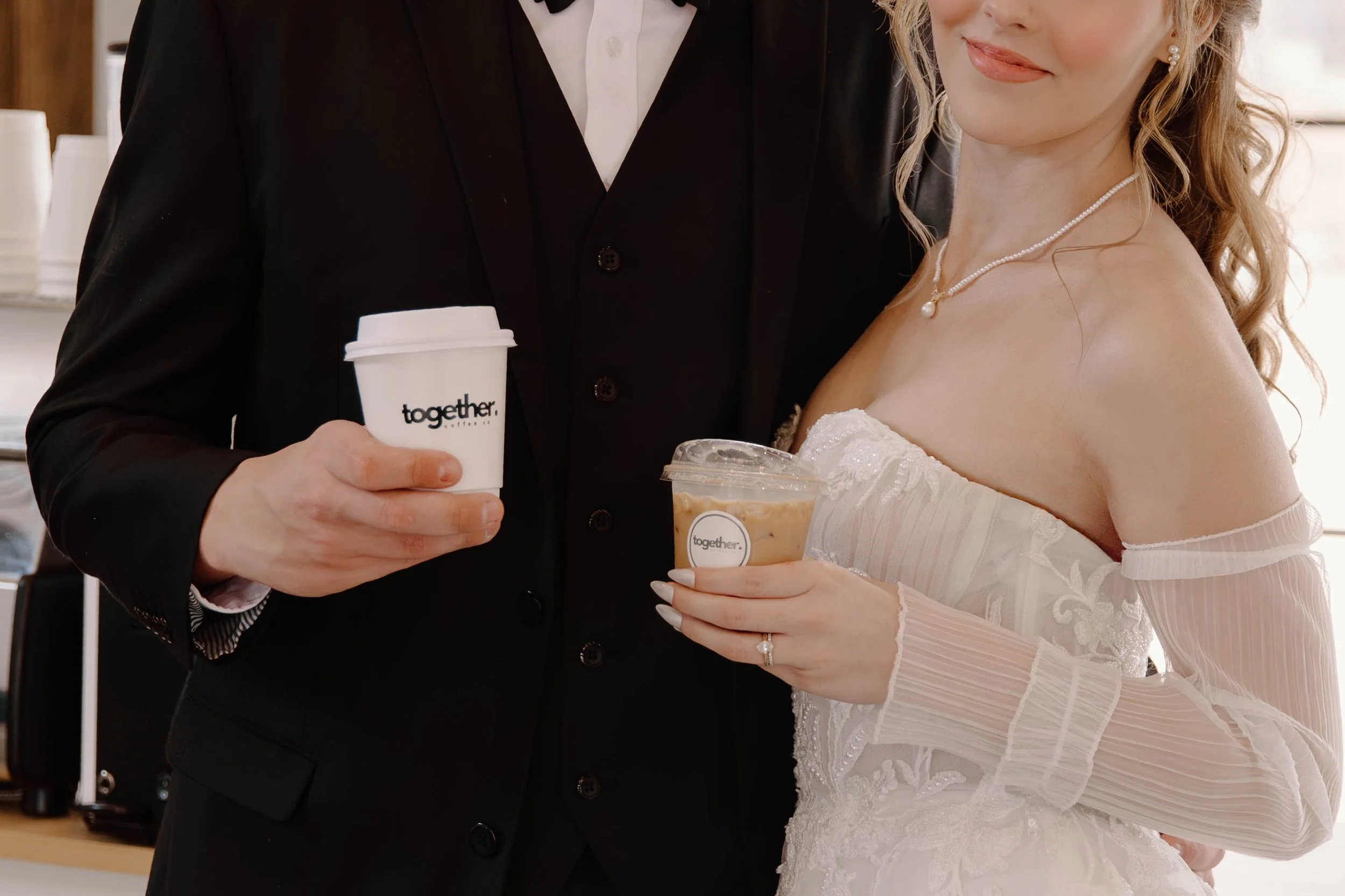 A smiling woman with dark wavy hair wearing a gray cap with the words 'YOUTH DEPT', a yellow T-shirt that says 'together.', and green pants, holding a glass of iced coffee, standing behind a coffee counter.