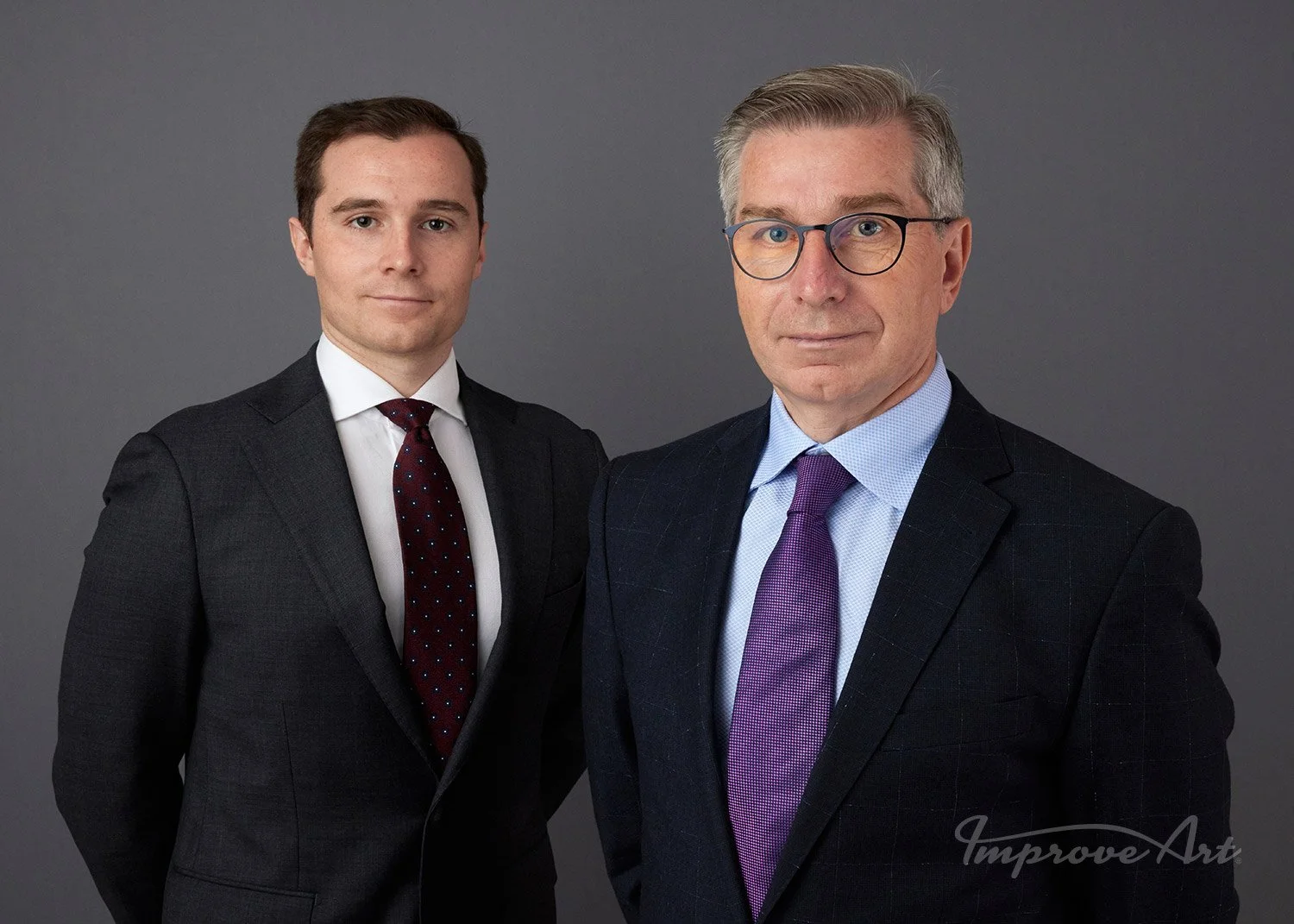 Two men in suits posing against a gray background.
