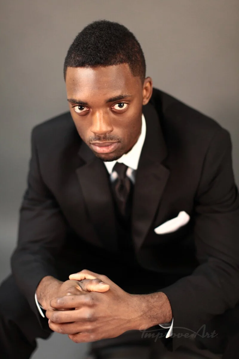 A young man in a black suit and tie, sitting with hands clasped, looking directly at the camera against a plain gray background.