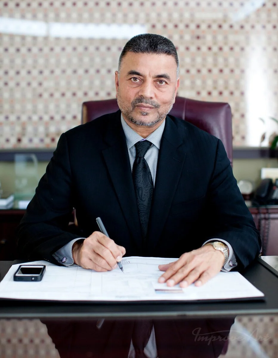 A middle-aged man in a black suit and tie sits at an office desk, holding a pen and looking at the camera. There is a smartphone on the desk and a large open folder with documents. He has short, gray hair and a beard, with a background of a patterned