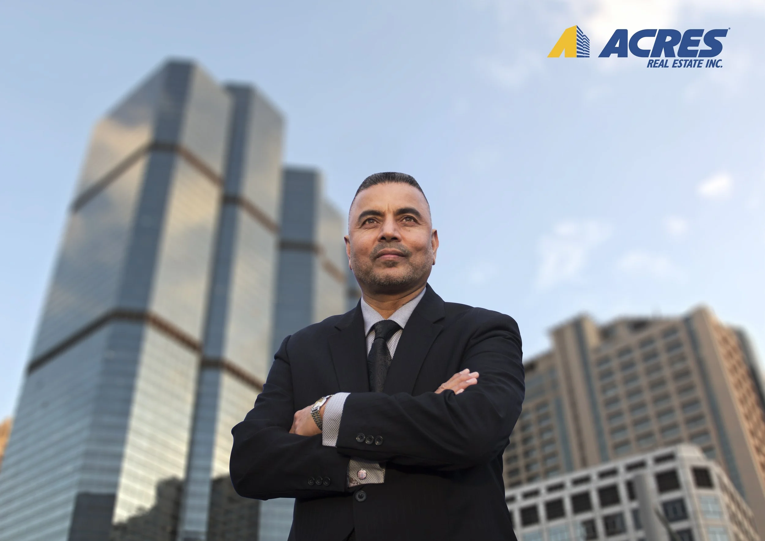 A confident man in a black suit with arms crossed in front of tall modern office buildings with the Acres Real Estate Inc. logo in the top right corner.