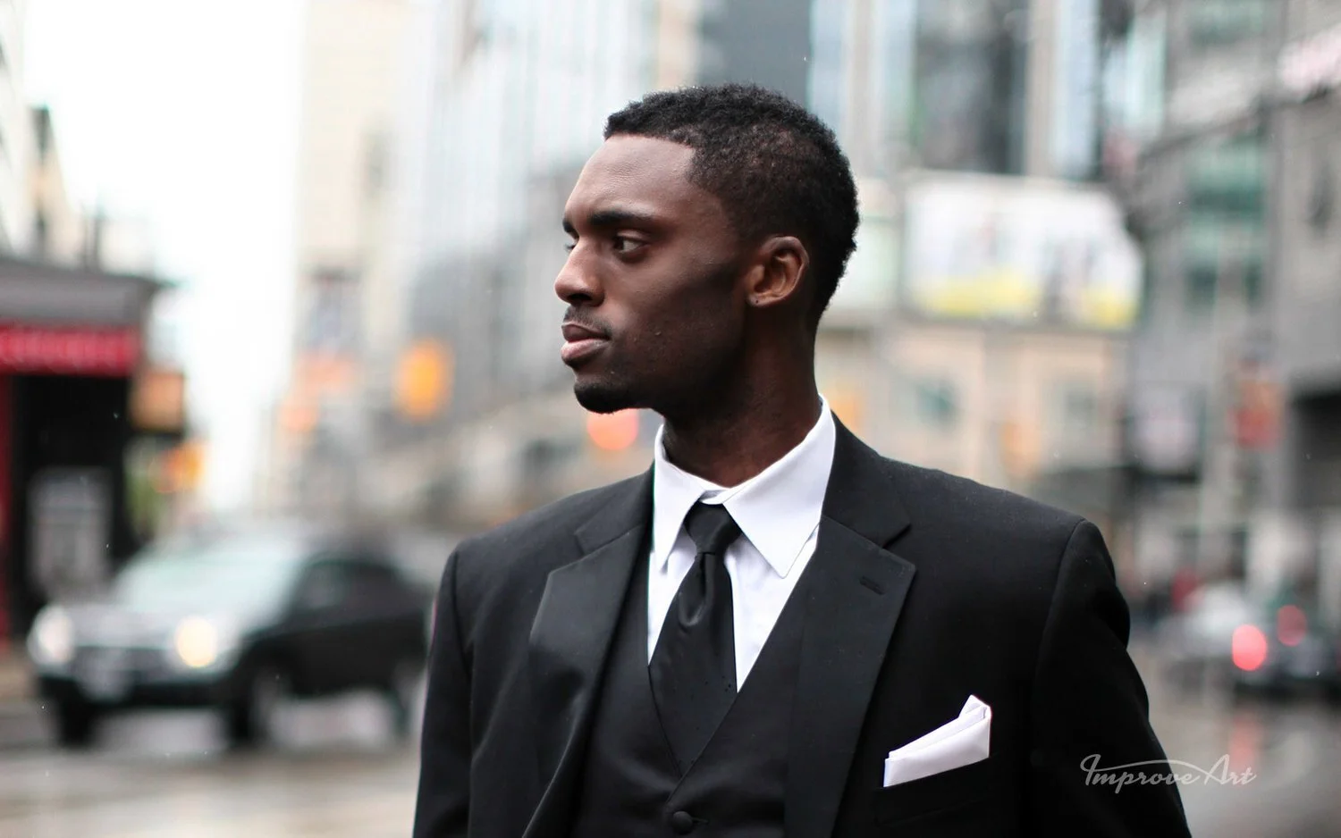 A man in a black suit and white shirt standing on a city street, looking to the side. Blurred cityscape with cars and buildings in the background.