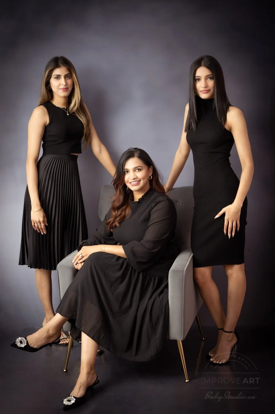 Three women in black dresses posing in a photo studio against a dark grey background, one seated in an armchair and two standing on either side.