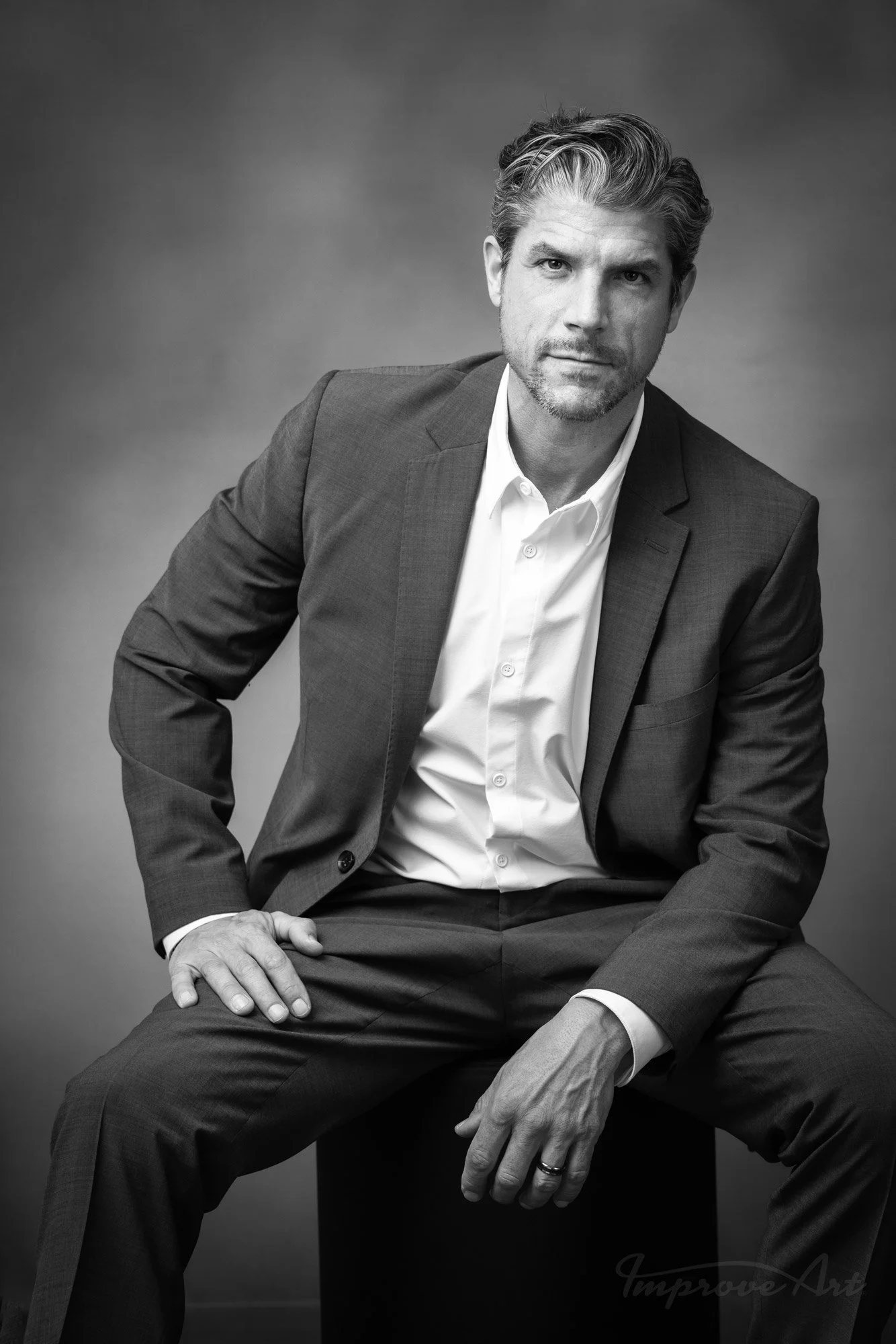 Black and white portrait of a man in a suit, seated with one hand resting on his knee and the other on his thigh, looking confidently at the camera.