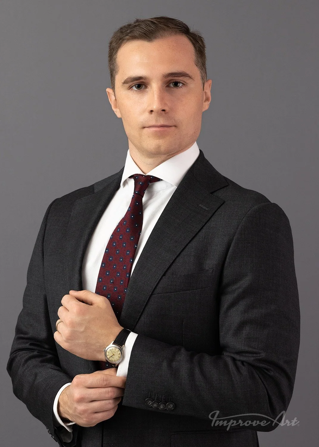 A young man in a dark suit, white shirt, and red polka dot tie, adjusting his shirt cuff, standing against a gray background.