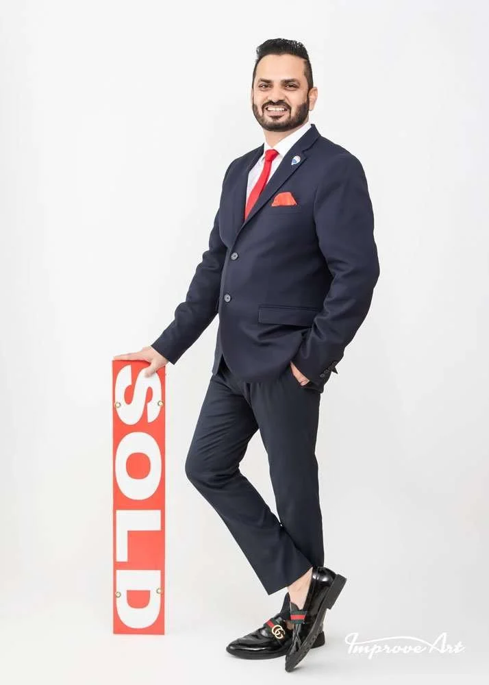 A man in a navy suit with a red tie and pocket square, smiling and standing against a plain white background. He has one hand in his pocket and is holding a red sign that reads 'SOLD' with his other hand. He is wearing black loafers with a logo and n