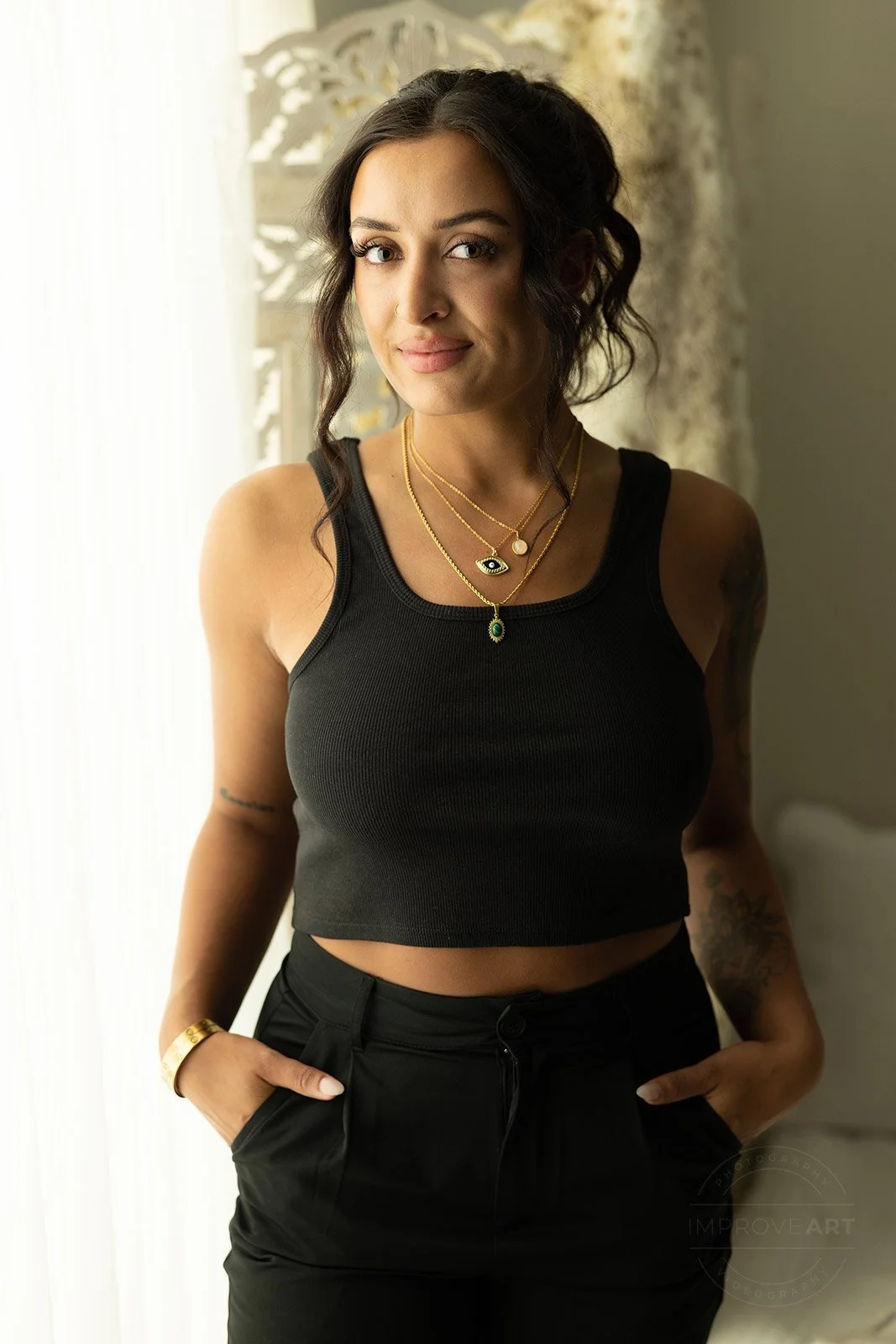 A young woman with dark wavy hair, wearing a black sleeveless crop top, black pants, and layered gold necklaces, standing indoors near a window with light coming through.