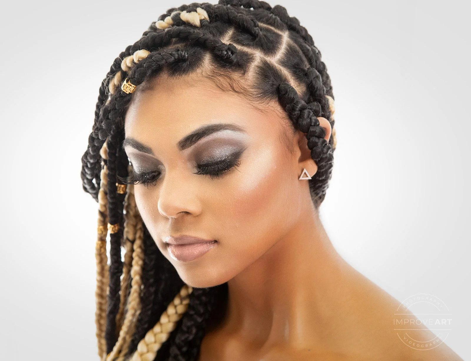 A woman with long, braided hair styled with black and blonde twists, adorned with gold beads and accessories, and wearing triangle-shaped earrings. She has makeup with smokey eyeshadow and a natural lip color, and her eyes are closed with a serene ex