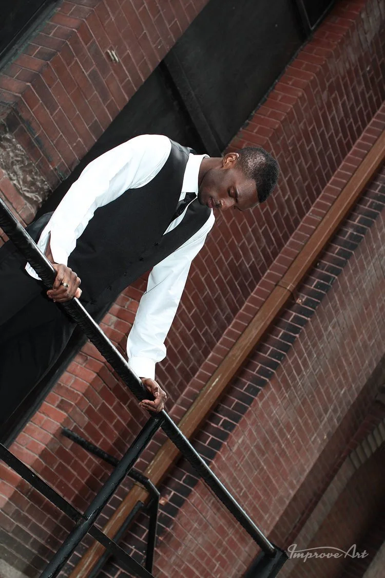 Young man in black vest, white dress shirt, and black tie standing on a staircase with black railing, looking down, against a brick wall background.
