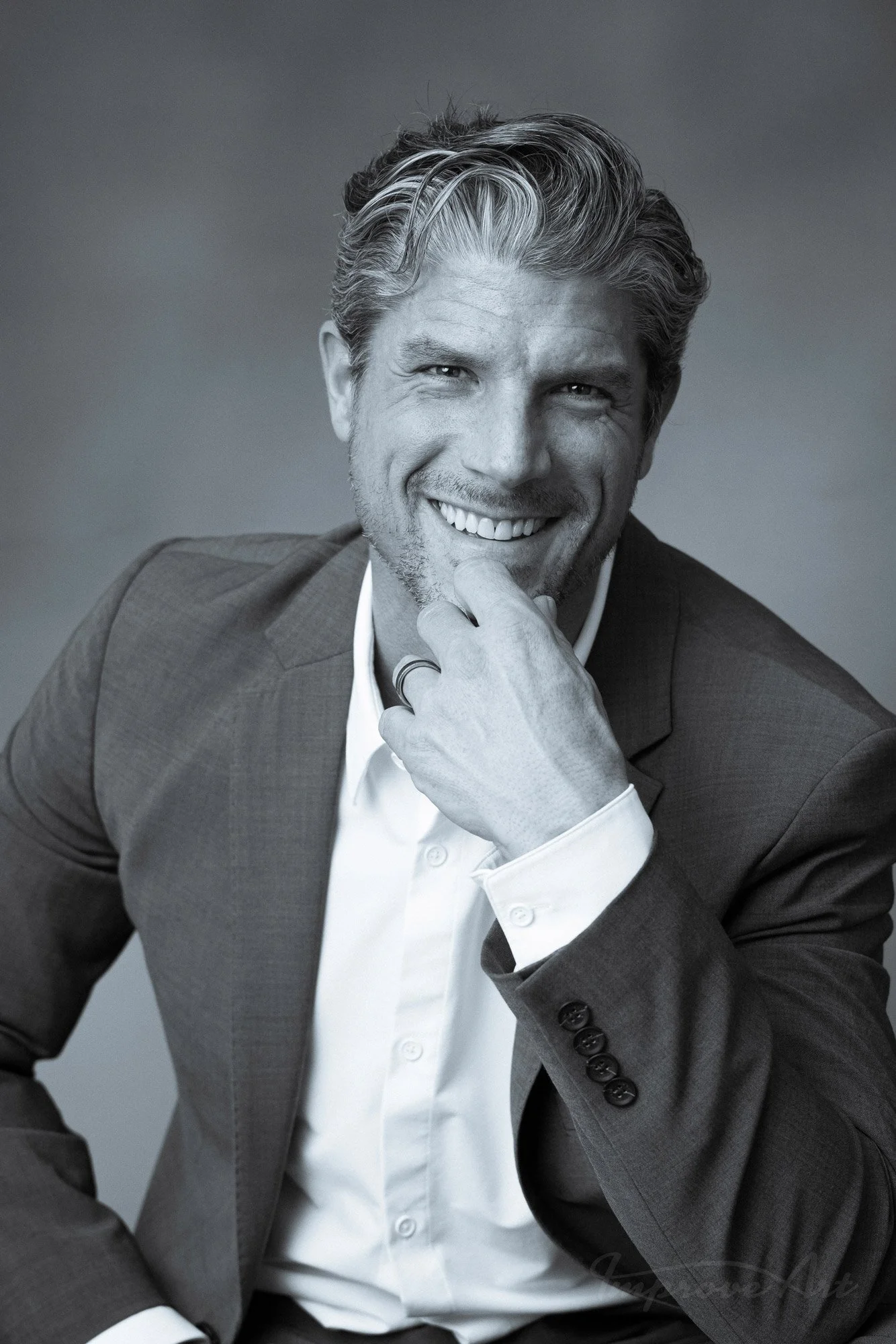 A black and white portrait of a smiling man with styled hair, wearing a suit and white dress shirt, resting his chin on his hand.