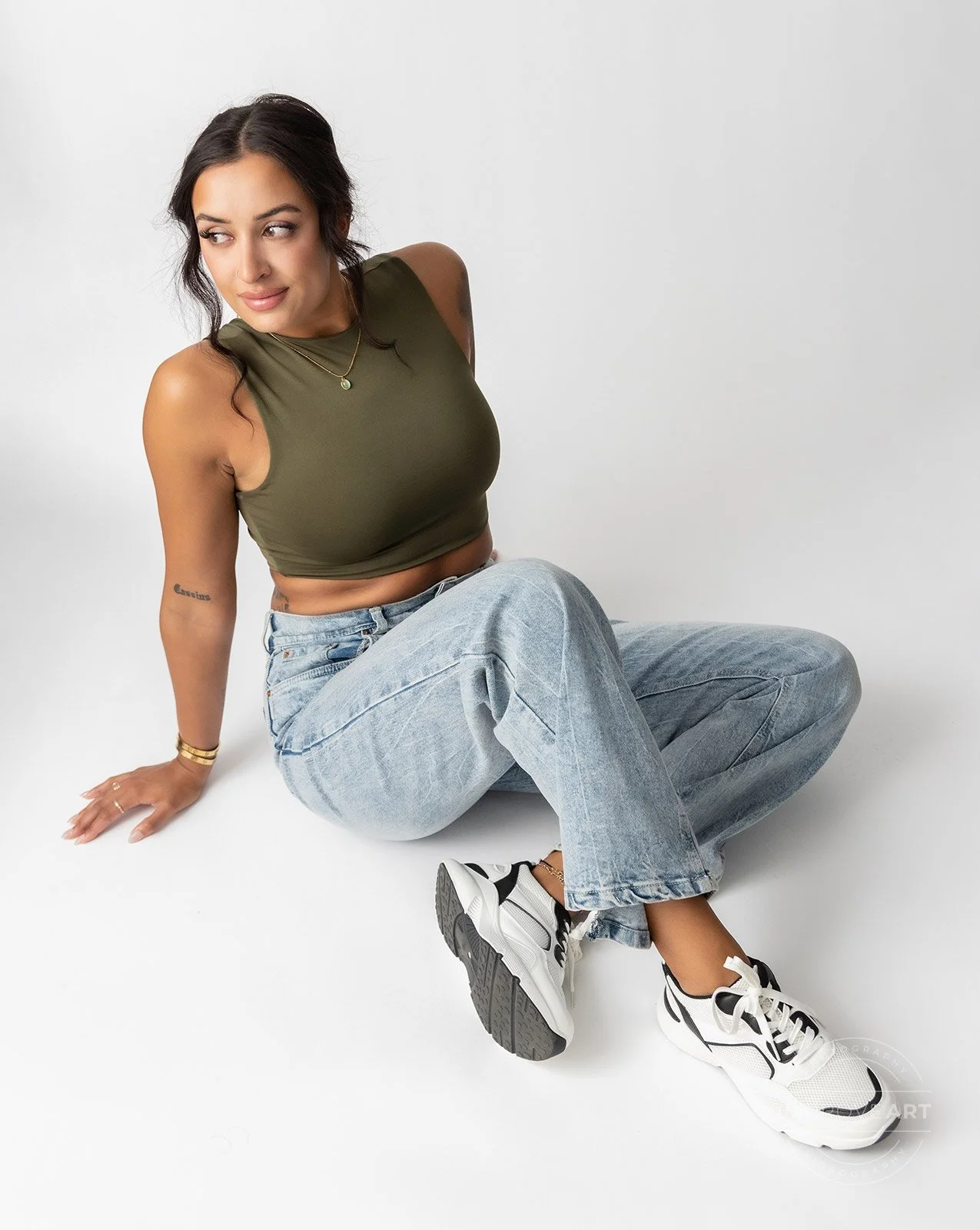A young woman with dark hair sitting on the floor, wearing a sleeveless olive green crop top, light blue baggy jeans, and white sneakers with black accents, against a plain white background.