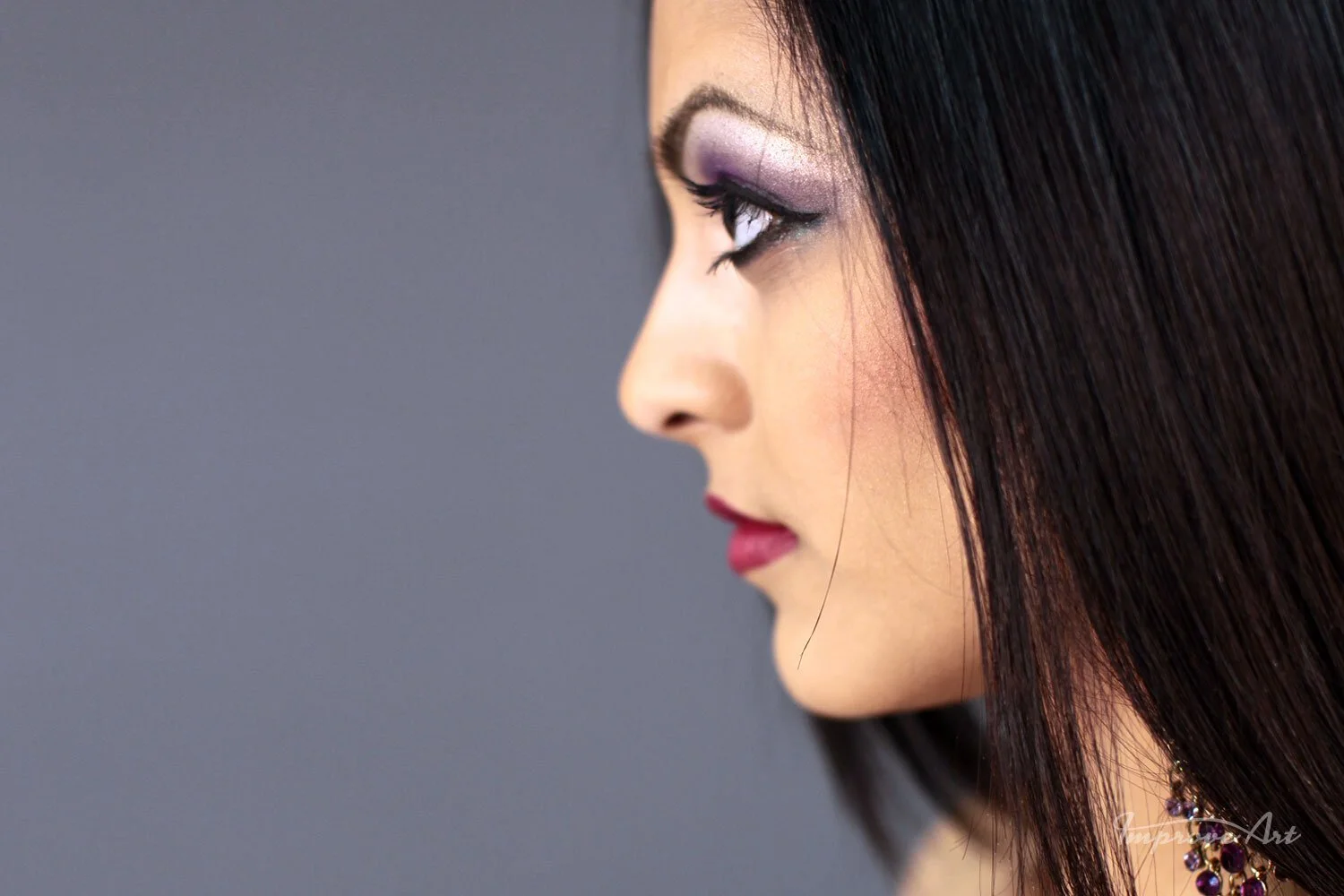 Close-up profile of a woman with dark hair, purple eyeshadow, and pink lipstick against a gray background.