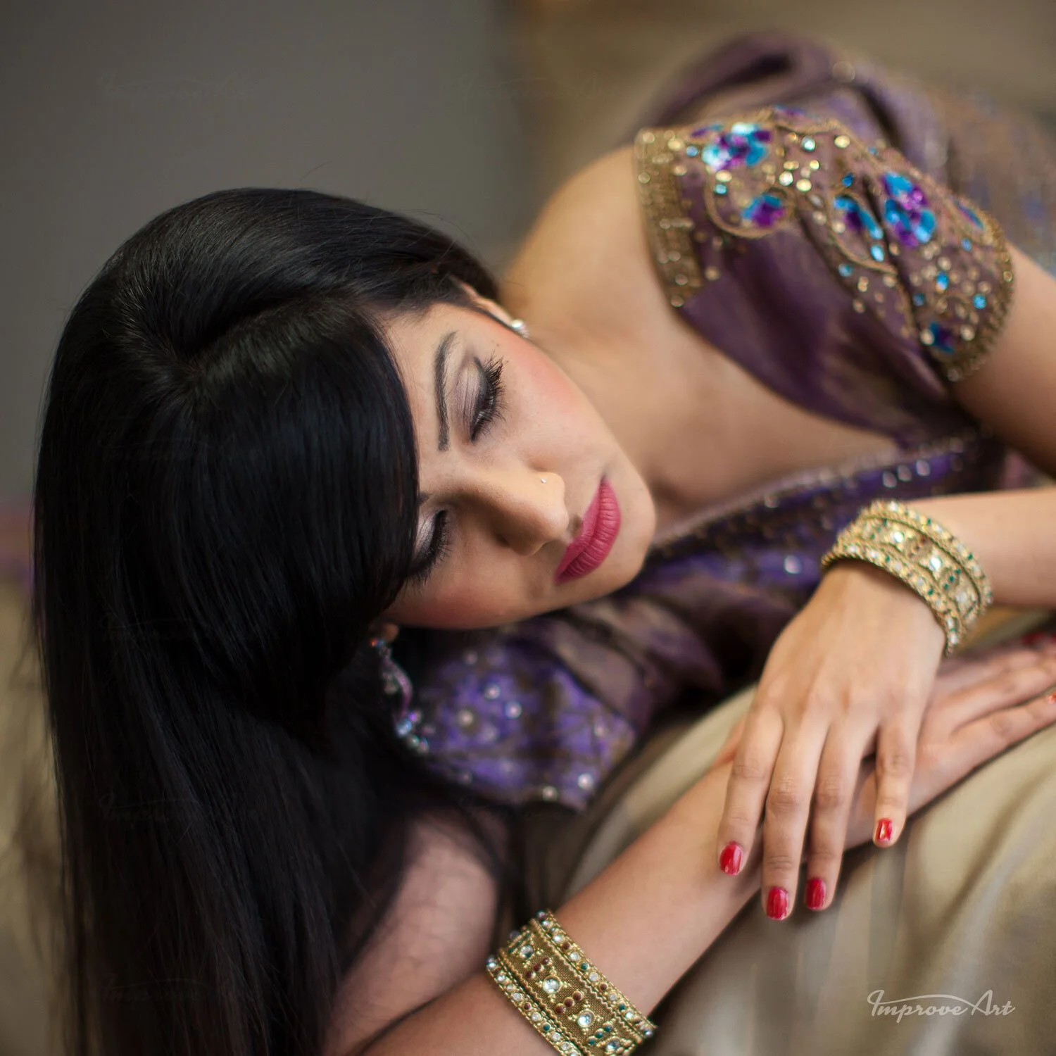 A woman with long black hair lying down, wearing purple traditional attire with gold and colorful embellishments, gold jewelry, and red nail polish, resting with eyes closed.