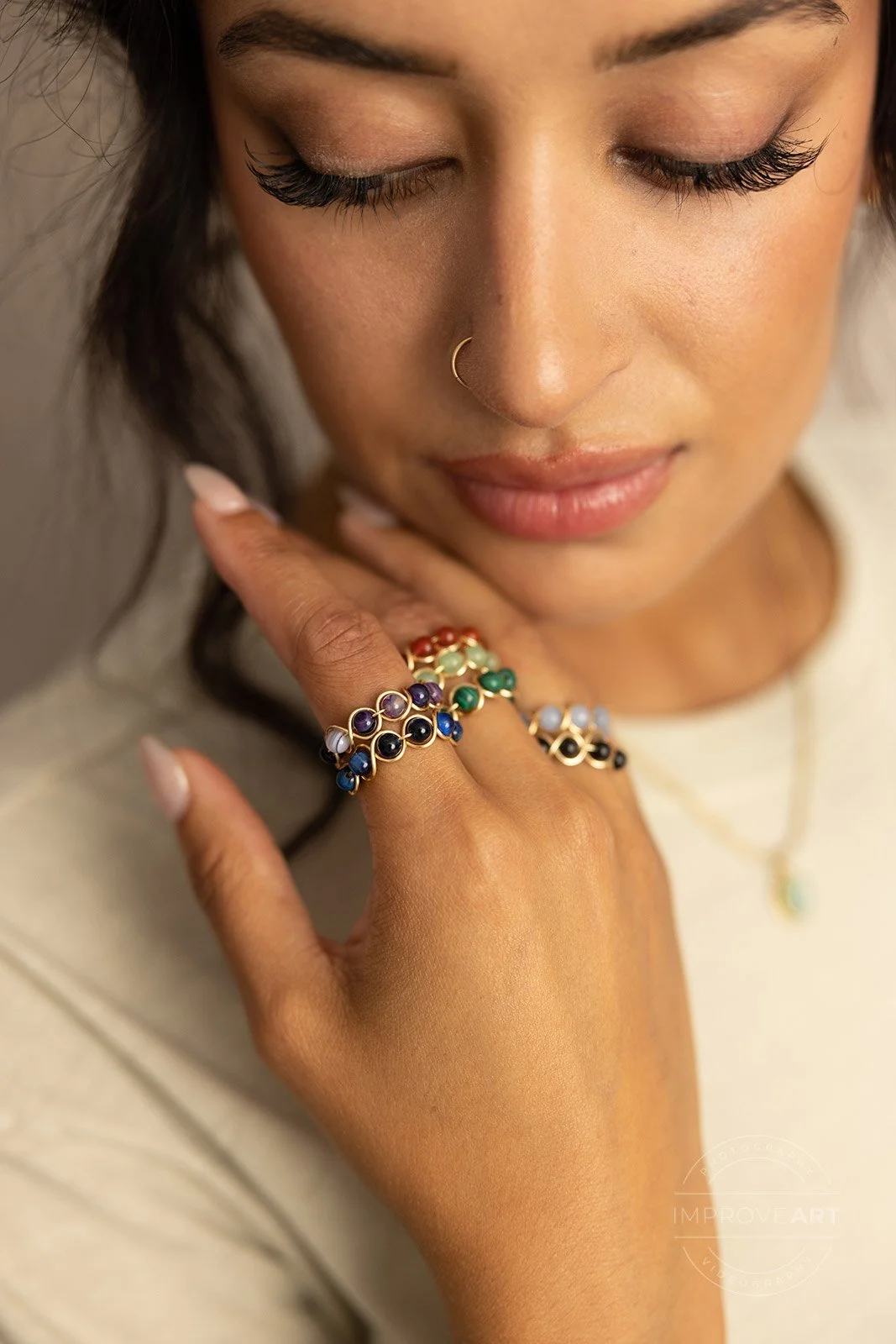 A woman with closed eyes, wearing a gold nose ring and multiple colorful rings on her fingers, touching her chin.