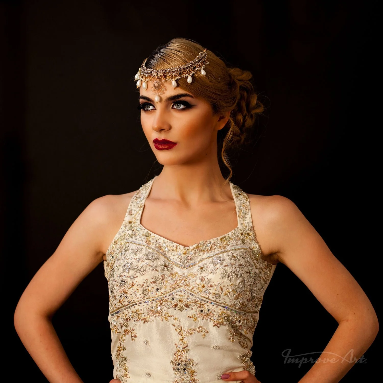 A woman with blonde hair styled in an updo, wearing jewelry including a headpiece with pearls, and a sleeveless, ornate, cream-colored dress with gold embroidery, posing against a dark background.