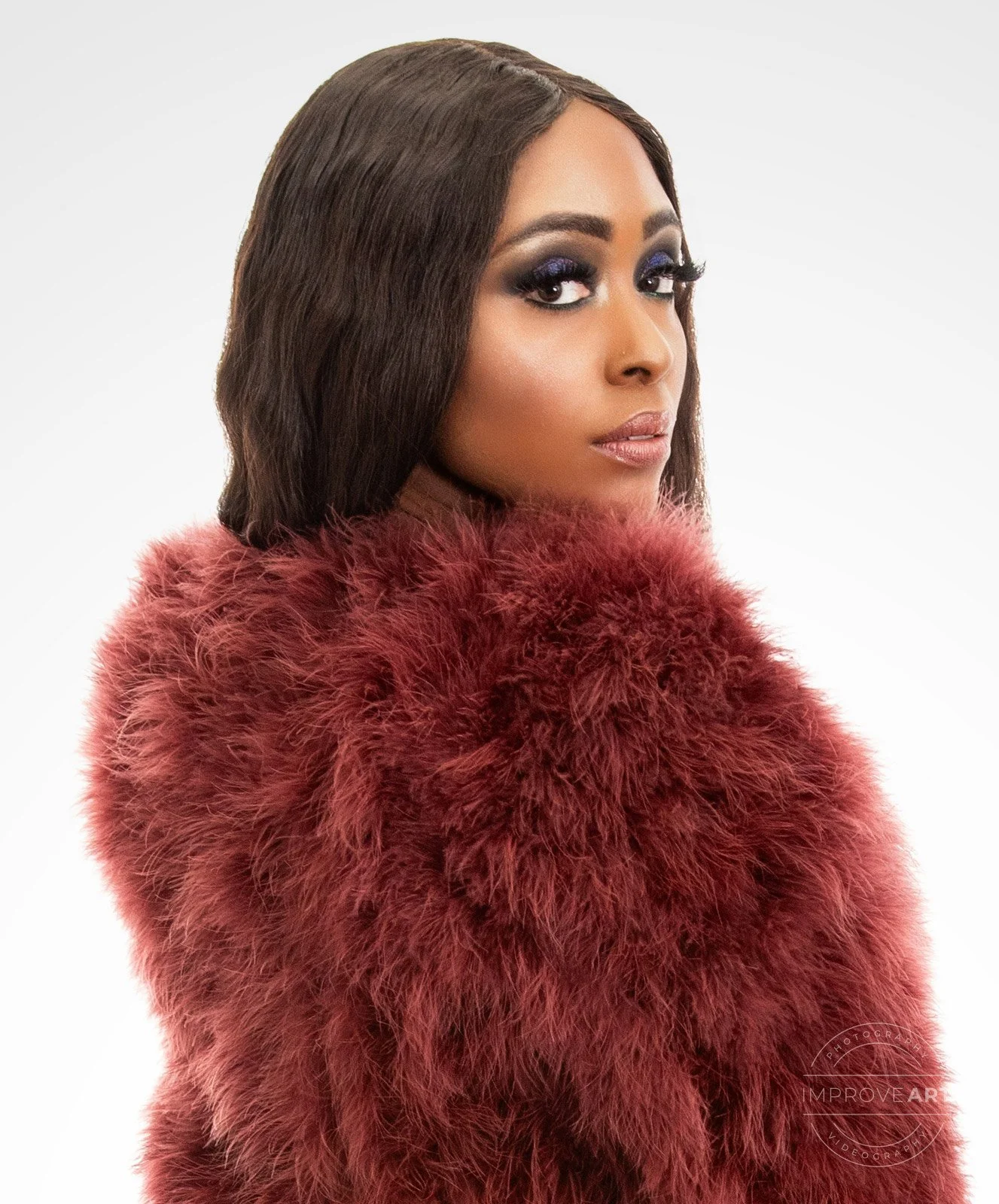 A woman with long dark hair and makeup posing in a red furry coat against a plain white background.