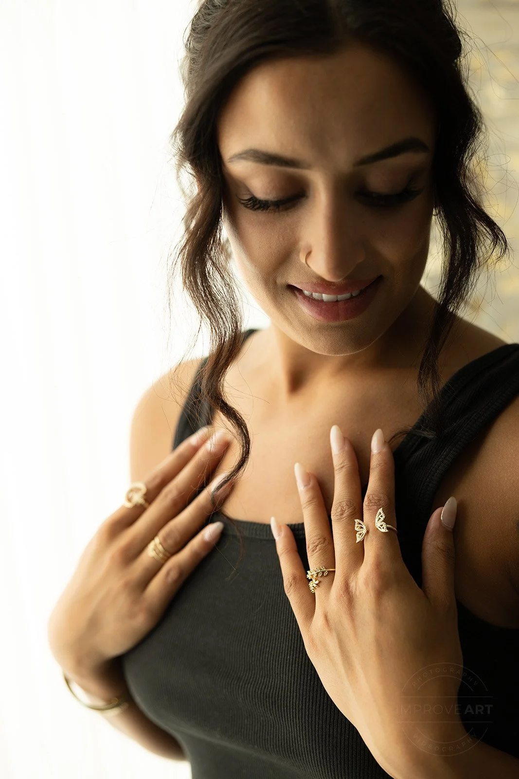 A woman with dark hair, wearing a black sleeveless top, smiling, with her eyes closed. She has rings on her fingers and is touching her chest with her hands.