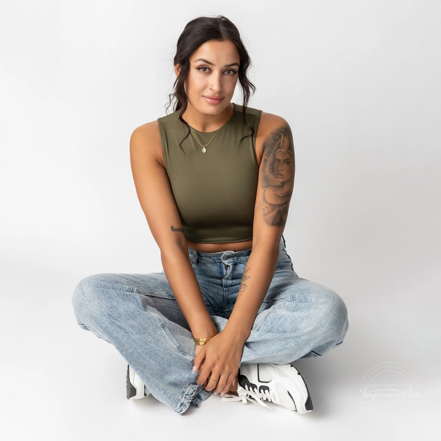 A young woman with tattoos, dark wavy hair, wearing a green sleeveless crop top, light-wash jeans, and white sneakers, sitting cross-legged against a plain white background