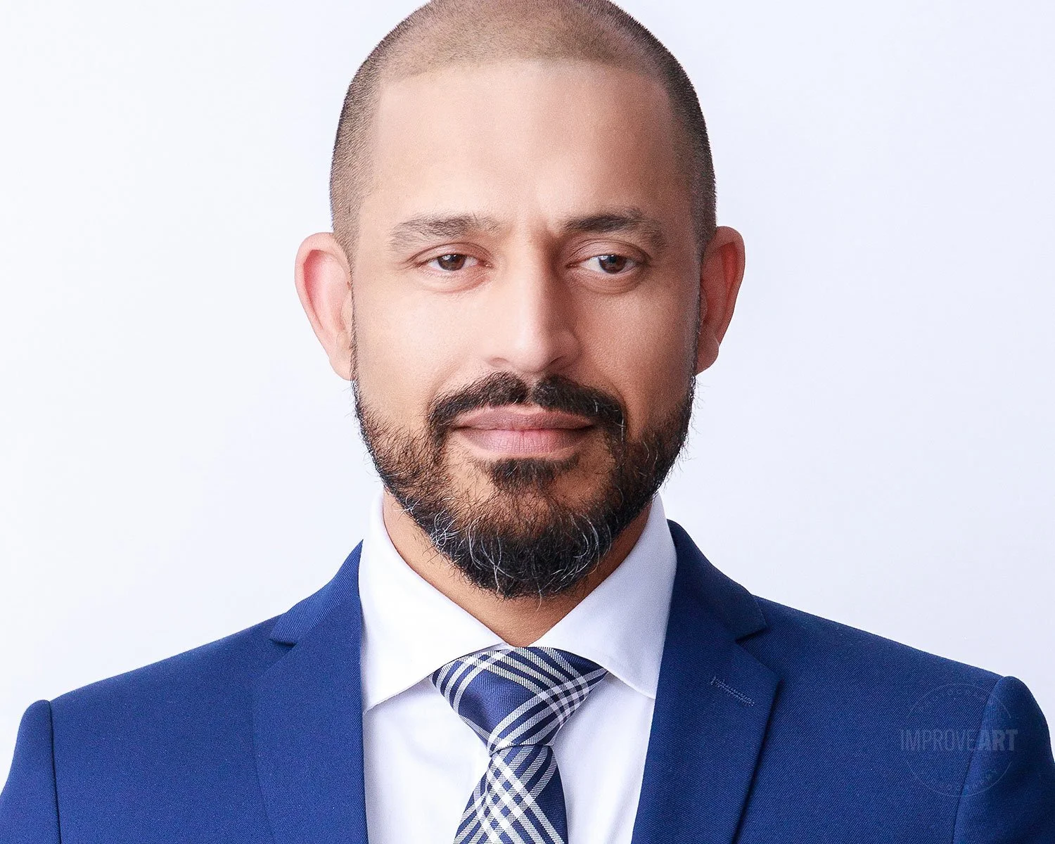 A man with a beard and mustache wearing a blue suit, white shirt, and striped tie, posing against a plain white background.