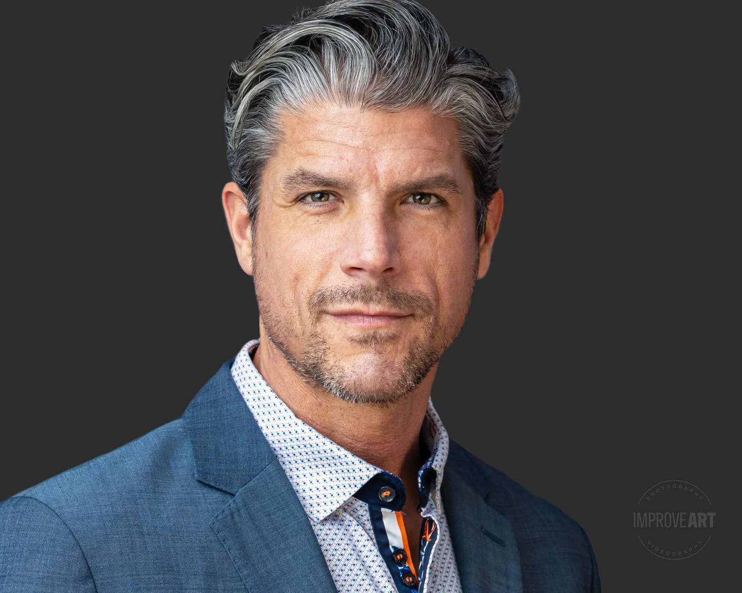 A middle-aged man with gray hair and blue eyes, wearing a blue suit and a white patterned shirt, looks into the camera against a dark background.
