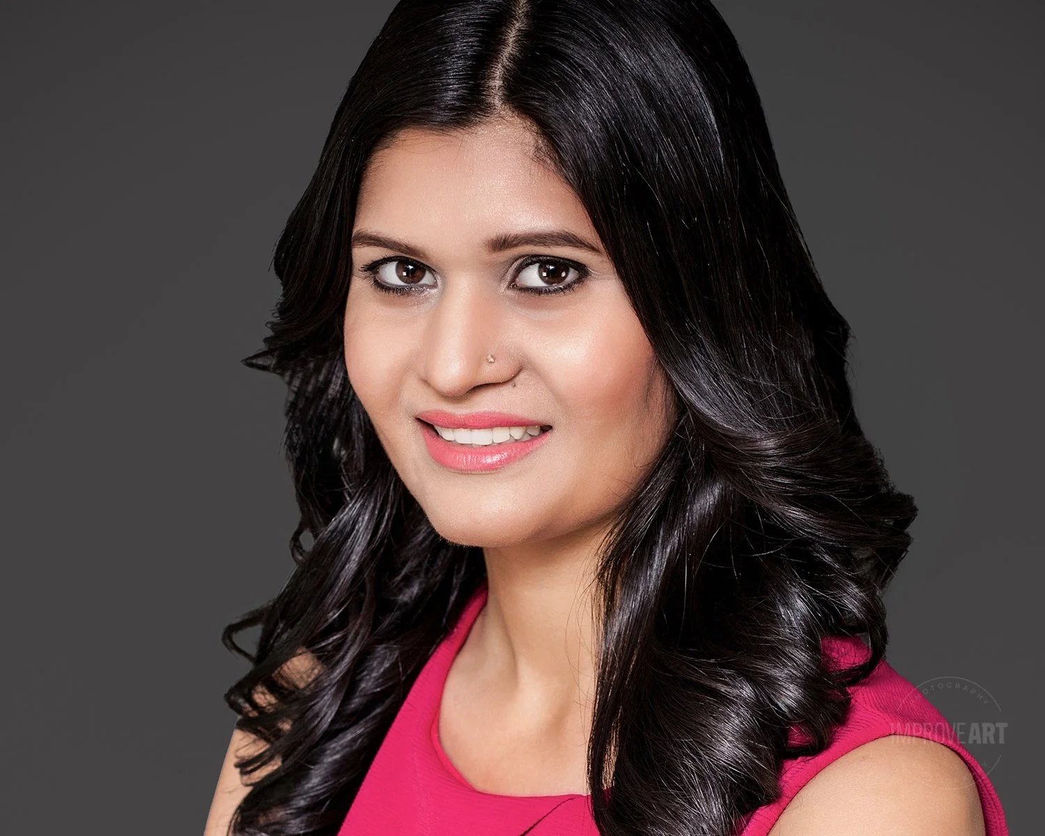A woman with long, dark, curly hair wearing a pink top, smiling against a dark gray background.