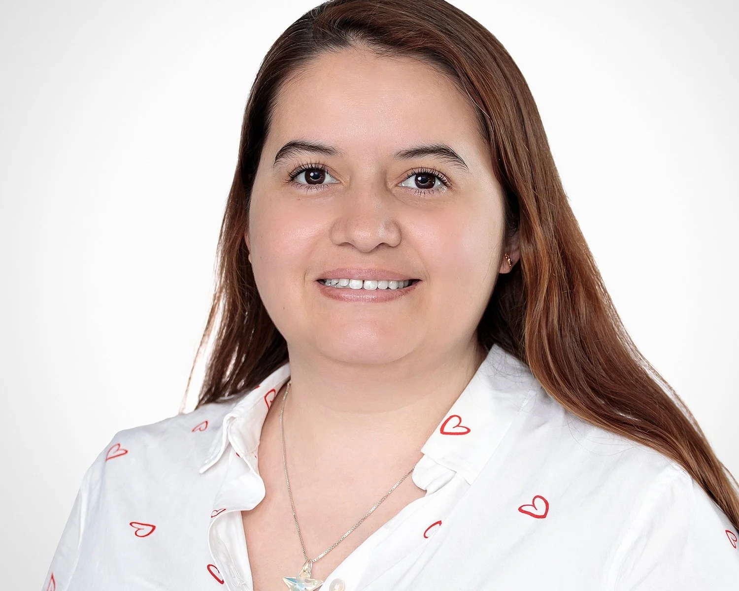 A woman with long brown hair wearing a white shirt with red hearts, smiling against a plain white background.