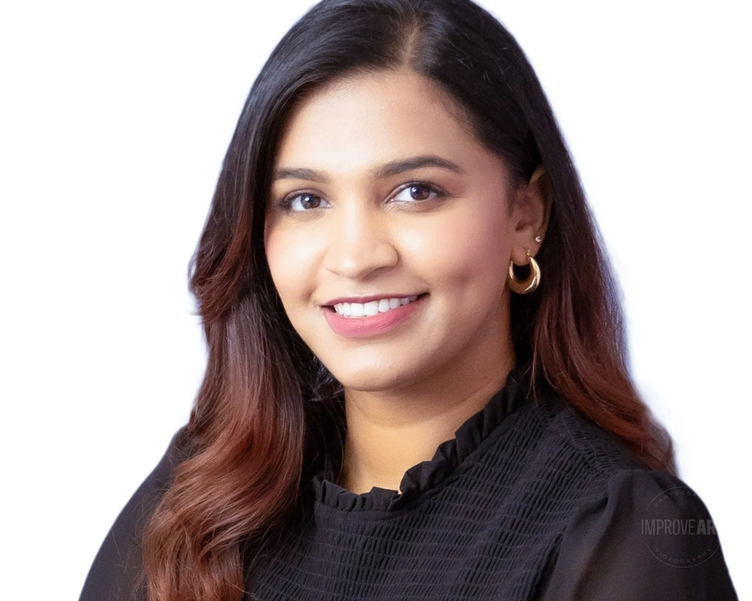 A professional headshot of a woman with long, wavy brown hair, wearing a black blouse with a high neck and ruffles, and gold hoop earrings. She is smiling against a plain white background.